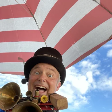 Gig Entertainer Ramp wearing a top hat and holding instruments under a red and white striped umbrella against a blue sky